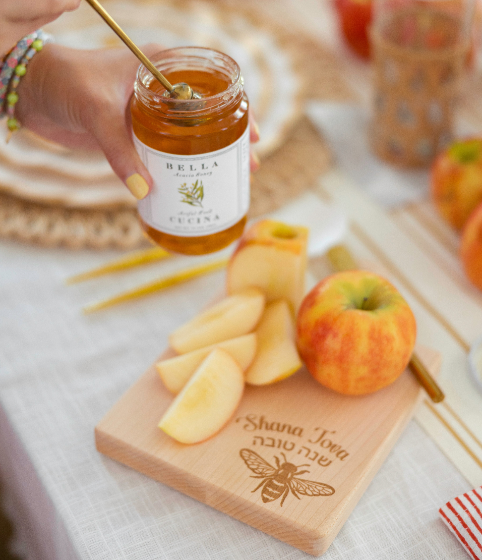 A hand holds a jar of honey with a spoon above sliced apples on a wooden board engraved with a bee and the words “Shana Tova” in Hebrew and English. An apple, Sophistiplate LLC’s Rosh Tablescape for 16 Guests: Golden Apple heavy duty paper plates, and additional festive foods complete the Rosh Hashanah table setting.