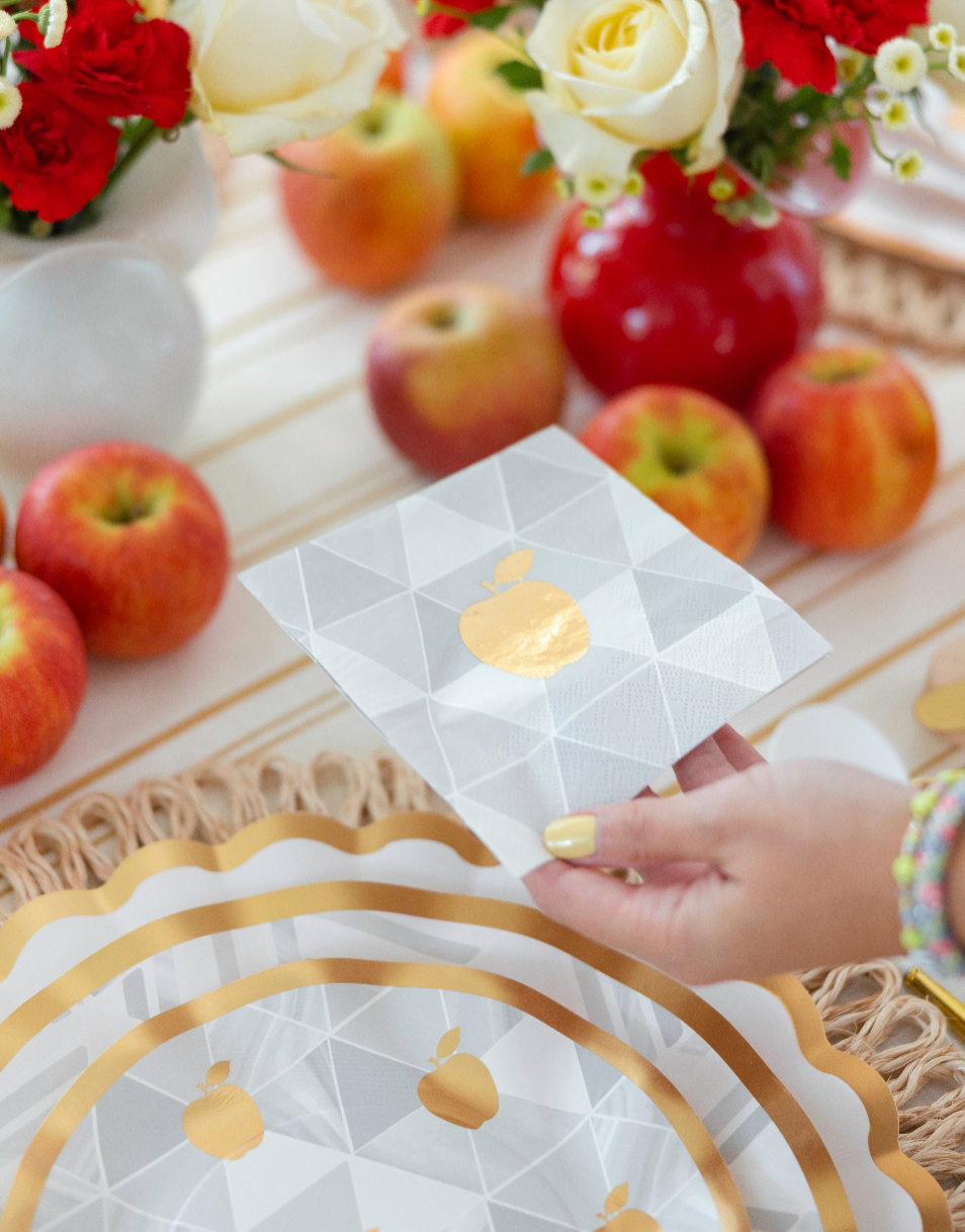 A hand holds a white napkin featuring a gold apple design from the Sophistiplate LLC “Rosh Tablescape for 16 Guests: Golden Apple” collection, surrounded by apples, floral arrangements, and matching heavy duty paper plates with gold accents on a beautifully decorated Rosh Hashanah table setting.