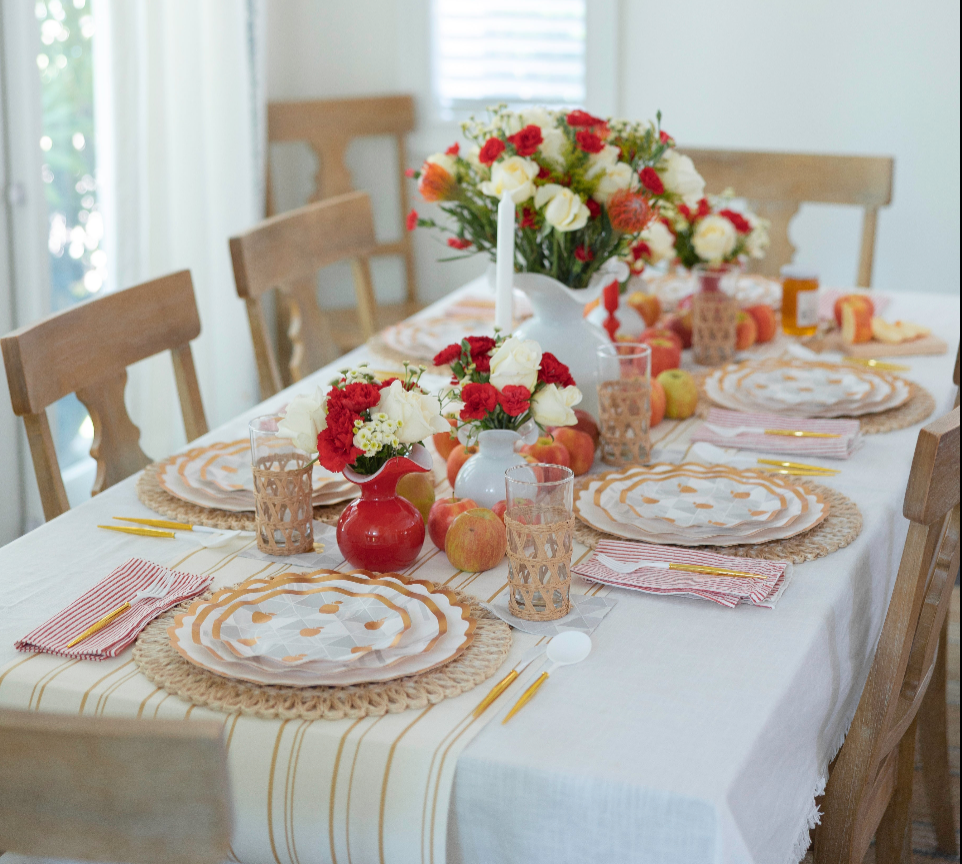 The Rosh Tablescape for 16 Guests: Golden Apple by Sophistiplate LLC features a dining table set with woven placemats, patterned plates inspired by Rosh Hashanah traditions, striped napkins, glasses, yellow utensils, and floral centerpieces accented with apples. Four wooden chairs are arranged around the display.