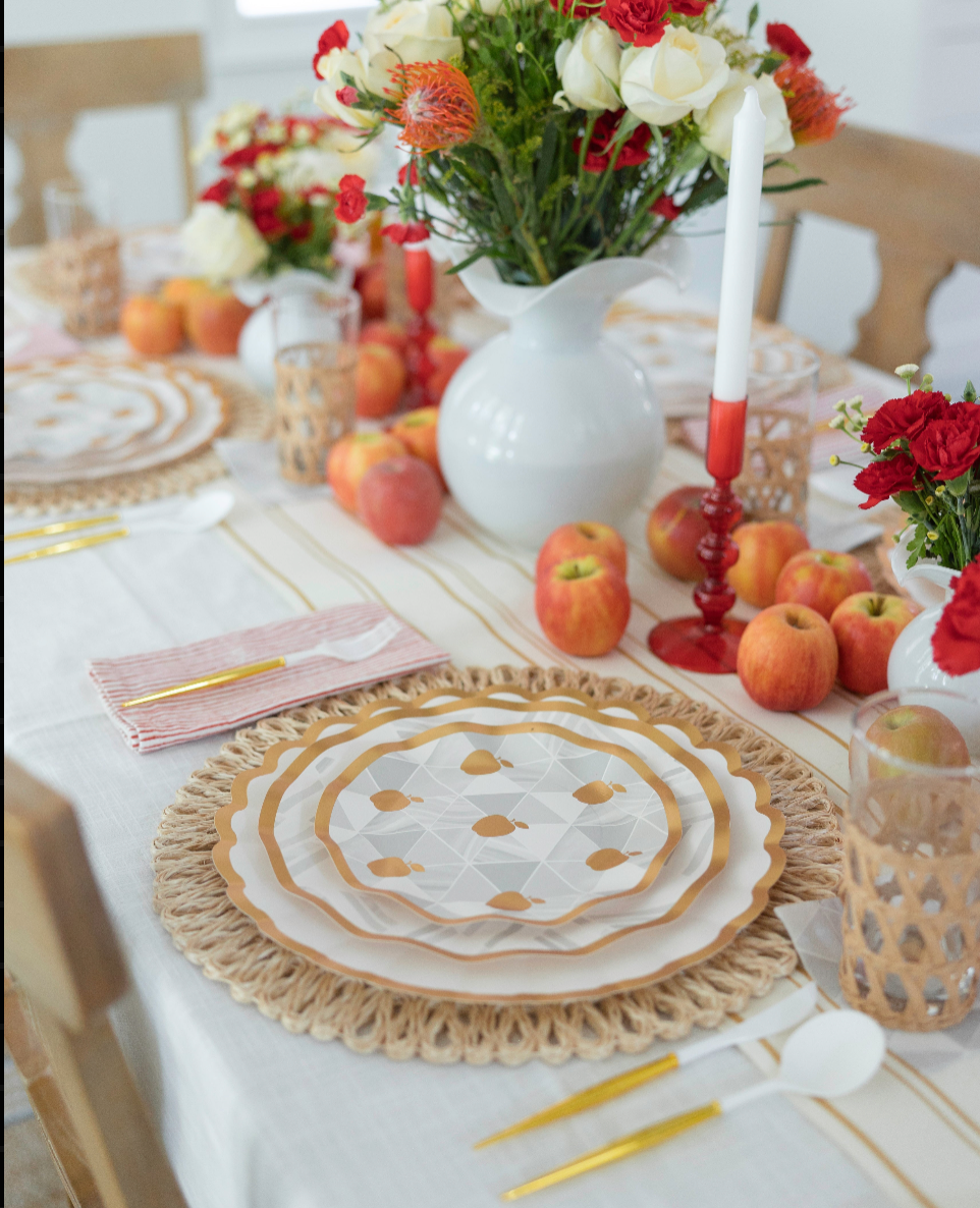 The Sophistiplate LLC Rosh Tablescape for 16 Guests: Golden Apple features heavy duty paper plates, gold cutlery, red-striped napkins, apples, candles, and floral centerpieces with white and red flowers in vases to create a festive Rosh Hashanah table setting.