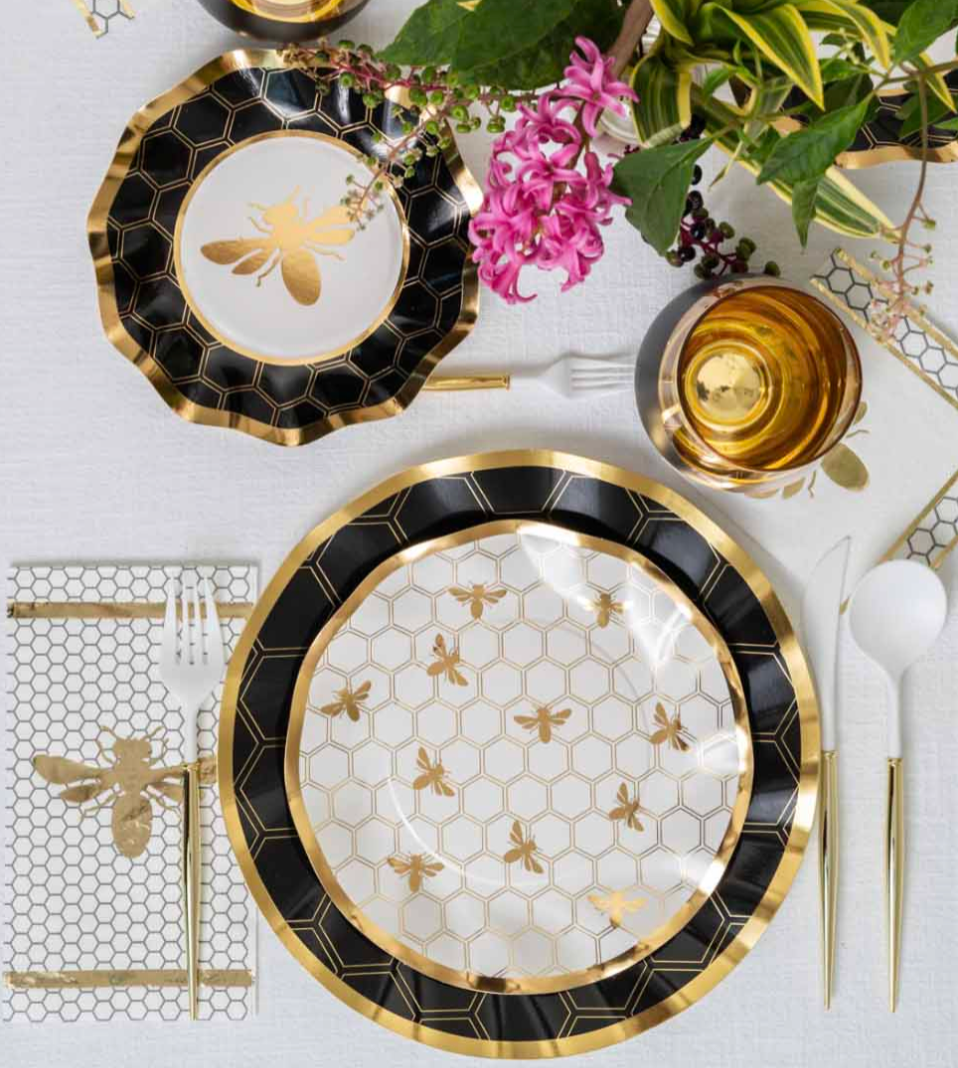 Elevate your Rosh Hashanah table with the Sophistiplate Rosh Tablescape for 16 Guests: Honeybee—an elegant ensemble featuring designer disposable plates, cups, and napkins adorned with gold bee and honeycomb patterns, paired with gold utensils, a pink hyacinth flower, and green foliage atop a white tablecloth for a truly stylish celebration.