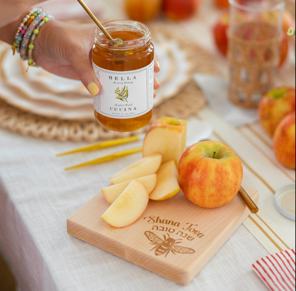A hand dips a spoon into a jar of honey near sliced apples on a wooden board engraved with “Shana Tova” and a bee, beautifully arranged on the Sophistiplate Rosh Tablescape for 16 Guests: Honeybee—an elegant designer disposable set perfect for your festive Rosh Hashanah celebration.