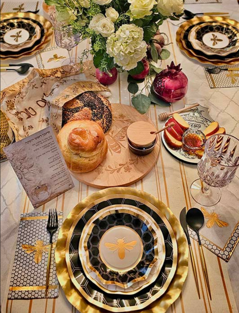 Set a festive Rosh Hashanah table with the Sophistiplate Rosh Tablescape for 16 Guests: Honeybee, showcasing designer disposable plates in elegant gold and black with charming bee motifs. The setting is complemented by challah bread, a honey jar, apples, pomegranate, wine glasses, and a floral centerpiece on a marble-patterned tablecloth.
