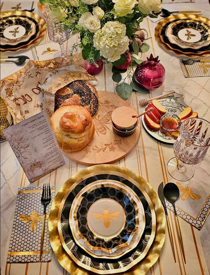 Set a festive Rosh Hashanah table with the Sophistiplate Rosh Tablescape for 16 Guests: Honeybee, showcasing designer disposable plates in elegant gold and black with charming bee motifs. The setting is complemented by challah bread, a honey jar, apples, pomegranate, wine glasses, and a floral centerpiece on a marble-patterned tablecloth.