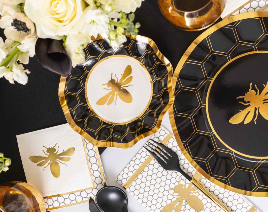 The Sophistiplate “Rosh Tablescape for 16 Guests: Honeybee” set features a chic black and gold Rosh Hashanah table setting, adorned with honeycomb patterns and bee designs on designer disposable plates, napkins, and cups, stylishly paired with black cutlery and accented by white flowers in a vase.
