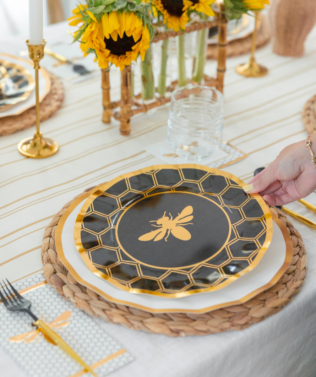 A hand sets down a Sophistiplate “Rosh Tablescape for 16 Guests: Honeybee” designer disposable plate, adorned with a gold bee and hexagon pattern, onto a woven placemat; sunflowers and gold utensils complete the elegant bee-themed Rosh Hashanah table setting.
