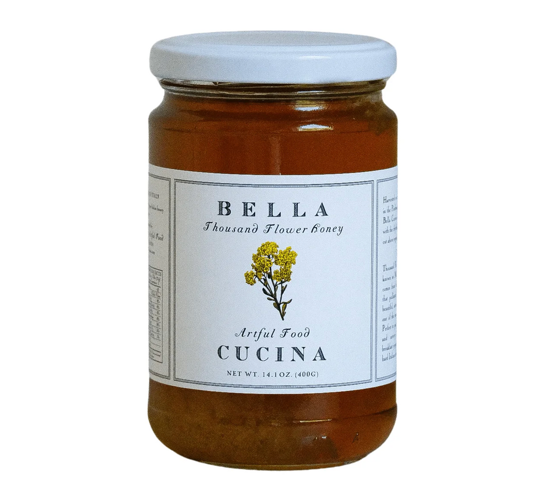 A glass jar of Bella Cucina Organic Thousand Flower Honey with a white lid and label, featuring a yellow flower illustration and text describing the product as Artful Food Cucina. Sourced from the wild blossoms of Piedmont, Italy, this biodynamic honey has a net weight of 14.1 oz (400g).