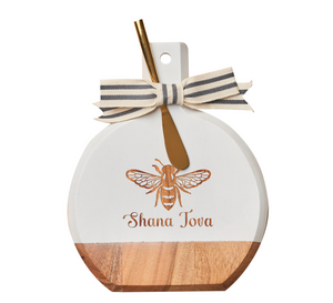 Celebrate Rosh Hashanah with the Maple Bevel Board w/ Spreader Knife | Shana Tova | 9.75 x 8″. This round acacia wood charcuterie board features a wooden and white design, beautifully engraved with a bee and “Shana Tova.” It comes topped with a striped bow and includes a gold spreader—perfect for festive gatherings.