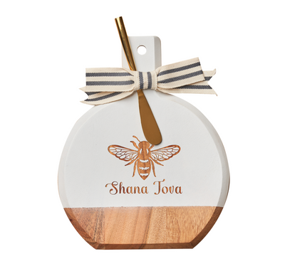 Celebrate Rosh Hashanah with the Maple Bevel Board w/ Spreader Knife | Shana Tova | 9.75 x 8″. This round acacia wood charcuterie board features a wooden and white design, beautifully engraved with a bee and “Shana Tova.” It comes topped with a striped bow and includes a gold spreader—perfect for festive gatherings.