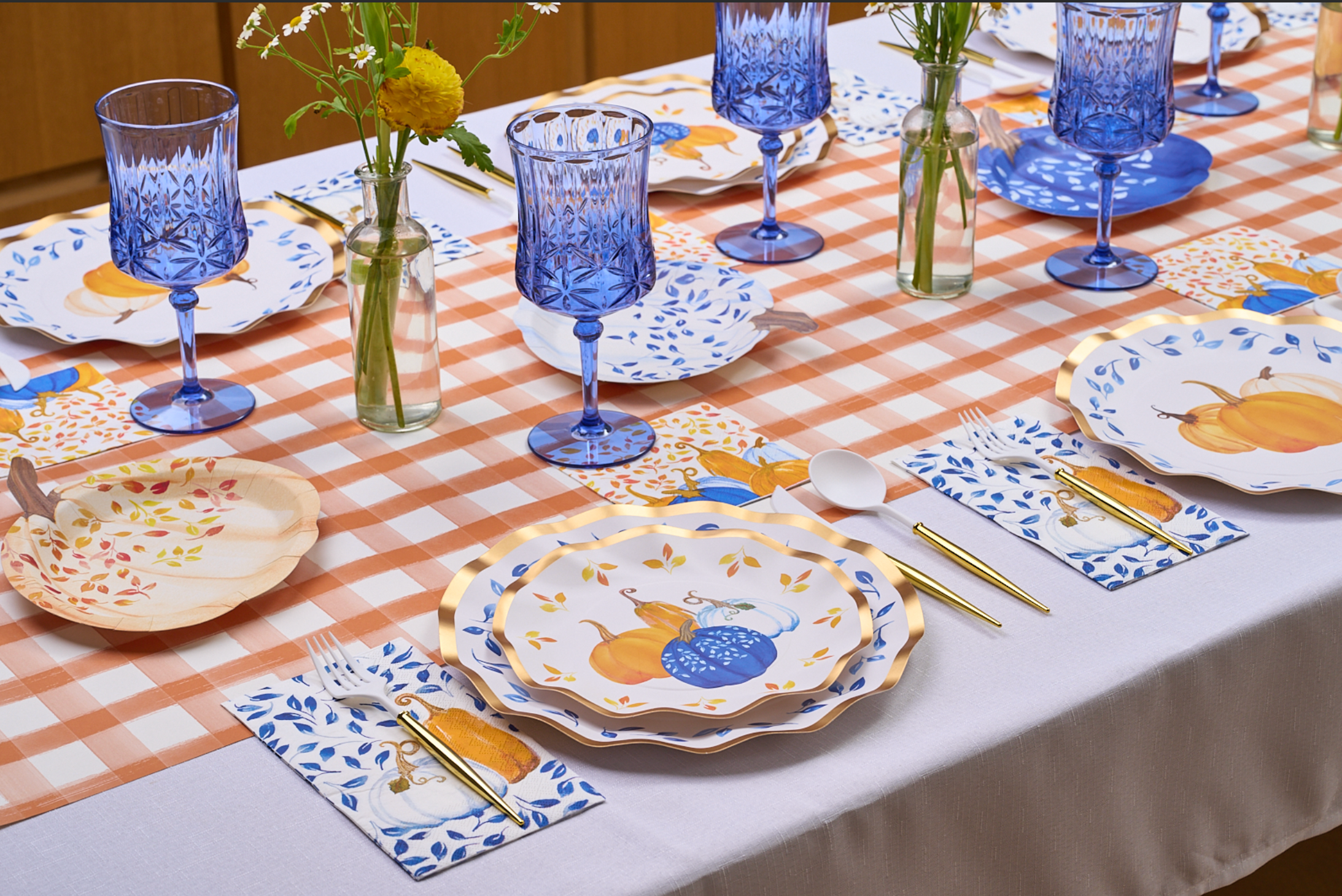 A table set with pumpkin-themed plates, blue goblets, gold cutlery, Sophistiplate’s Harvest Blues cocktail napkins, and small vases with yellow flowers on a checkered tablecloth—perfect for festive holiday gatherings.