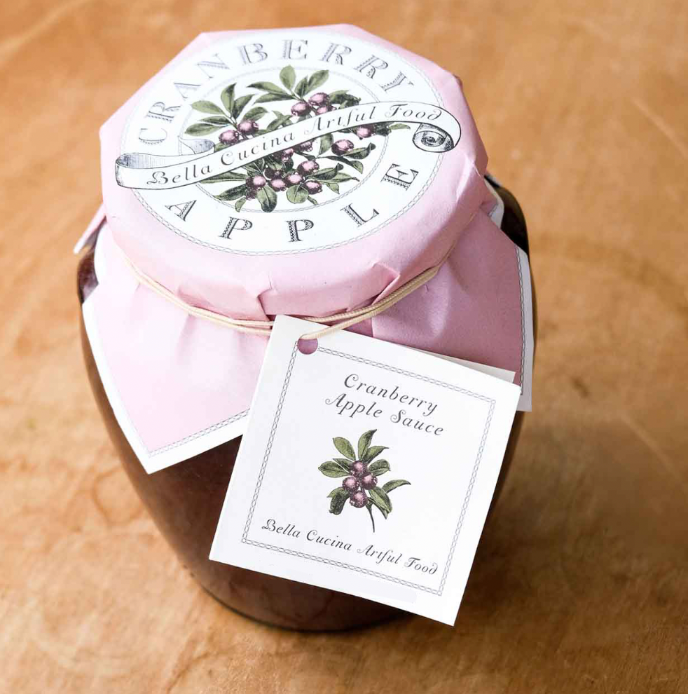 A glass jar of Bella Cucina Cranberry Apple Sauce features a pink lid, decorative watercolor-style apple and leaf accents on the label and tag, offering a sweet-tart sauce ideal as a Thanksgiving condiment.
