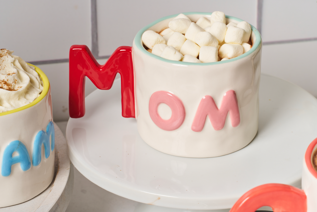 The Sophistiplate Ceramic Message Mug Mom, 14oz, featuring pink "MOM" letters and a red M-shaped handle, is filled with mini marshmallows and displayed on a white plate beside other colorful mugs.