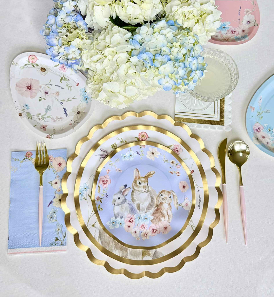 Create a charming spring table with Sophistiplate LLC's Ultimate Easter Tablescape for 16, featuring gold-rimmed scalloped plates with rabbits, pastel napkins, gold and pink cutlery, floral accents, and white and blue hydrangeas.