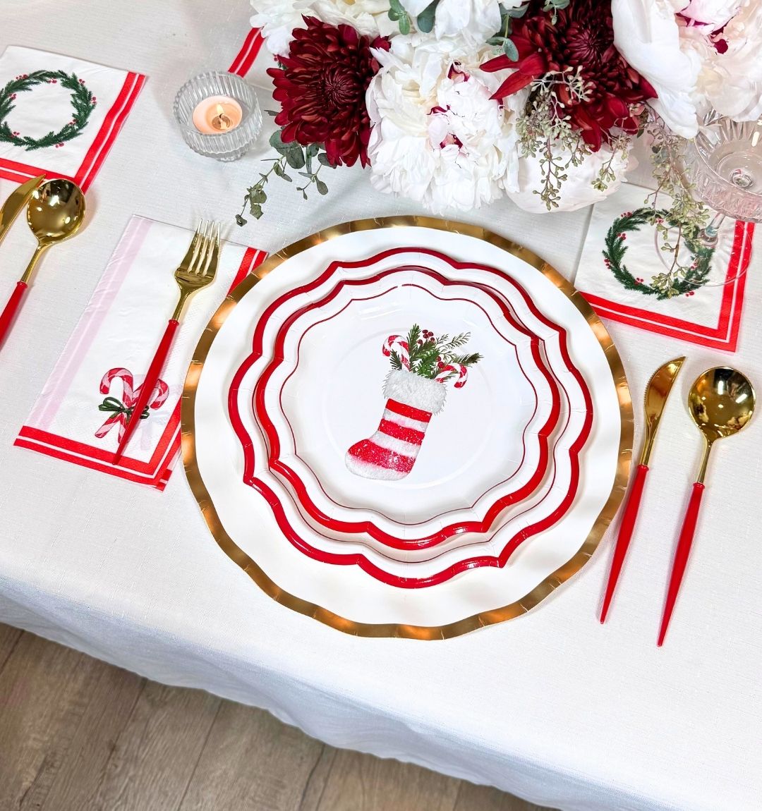 Create a festive holiday table with the Merry & Bright: Christmas Table for 16 set, featuring gold and red utensils, stocking-themed heavy duty plates, holiday napkins, and a red-and-white floral centerpiece on a white tablecloth.
