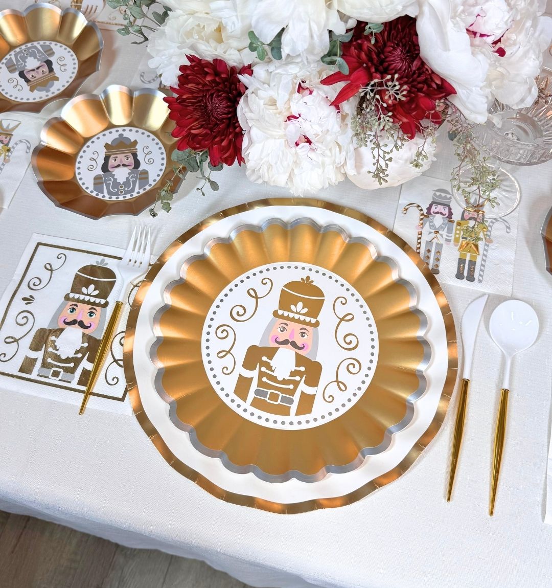 Sophistiplate Gilded Nutcracker: Christmas Table for 16 featuring nutcracker-design heavy duty paper plates, white and gold cutlery, and matching napkins, all arranged around a floral centerpiece of white and dark red flowers.