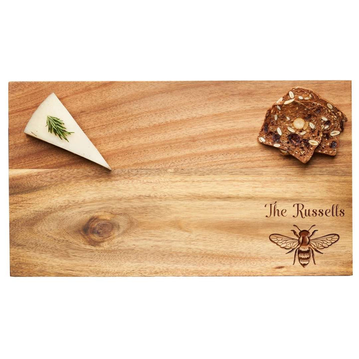 A wedge of herb cheese and seeded crackers sit on Maple’s 14" x 8" Acacia Wood Board, personalized with "The Russells" and a detailed honeybee illustration in the lower right corner.