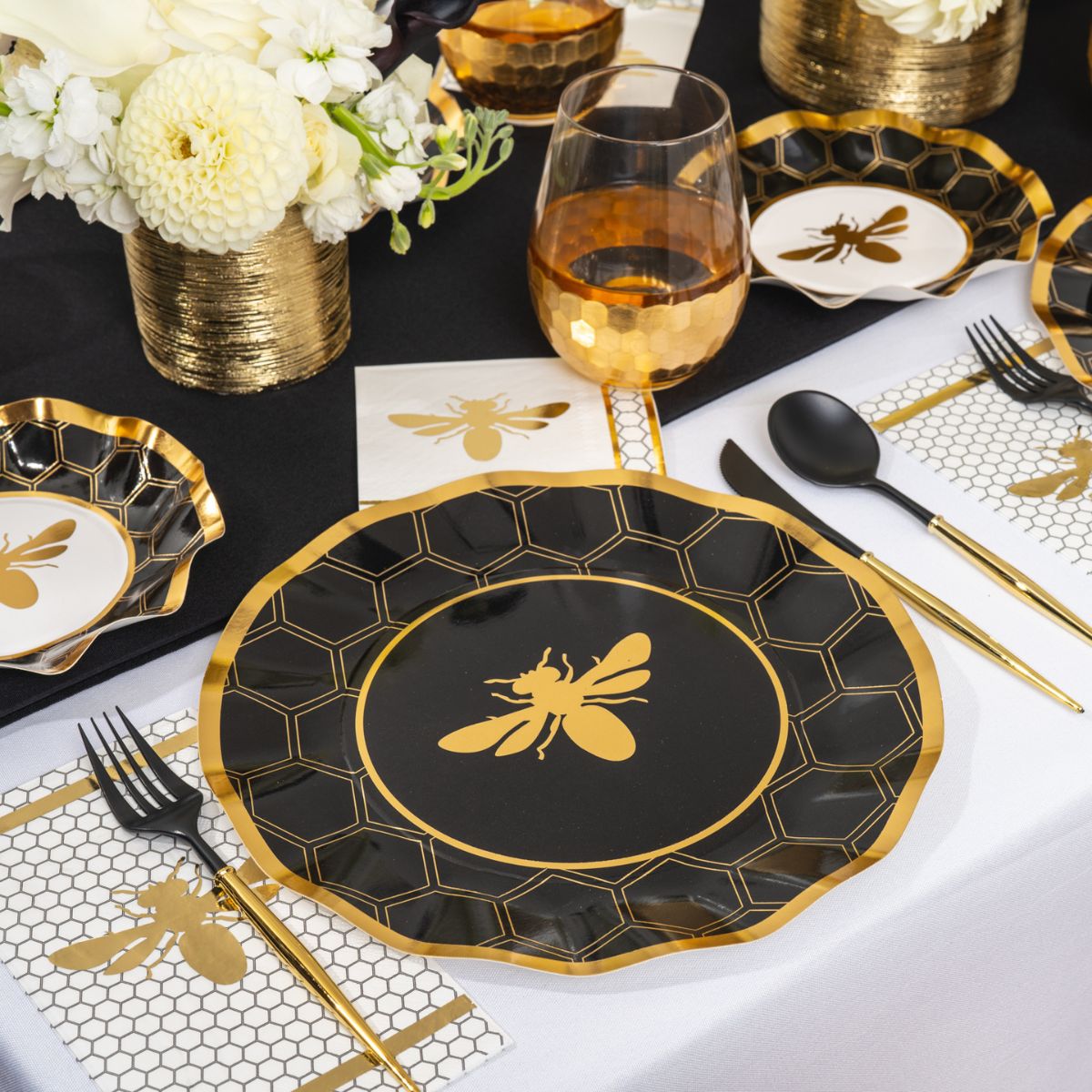 Create a sophisticated Rosh Hashanah table setting with the Sophistiplate “Rosh Tablescape for 16 Guests: Honeybee” set, featuring elegant black and gold designer disposable plates adorned with bee motifs, gold-rimmed glassware, chic black cutlery, and a white floral centerpiece. Honeycomb and bee designs accent the décor throughout, making it an ideal choice for a stylish holiday celebration.