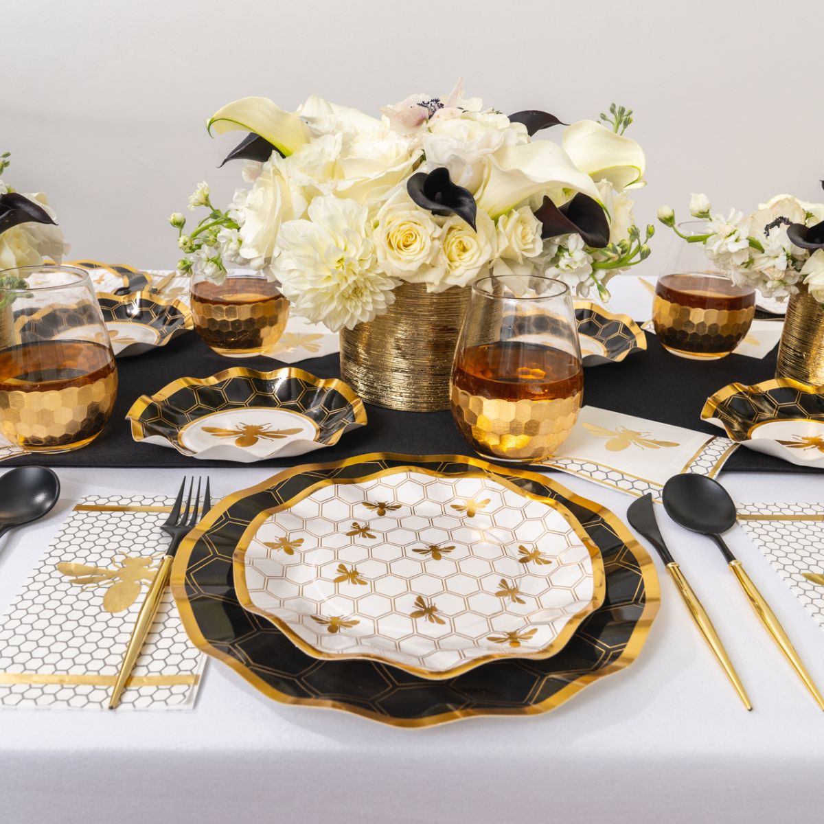 Create a stunning Rosh Hashanah table setting with the Sophistiplate Rosh Tablescape for 16 Guests: Honeybee, featuring elegant gold and black bee-themed heavy duty paper plates, matching black utensils, gold-rimmed glasses, and a beautiful centerpiece of white and yellow flowers in a gold vase.