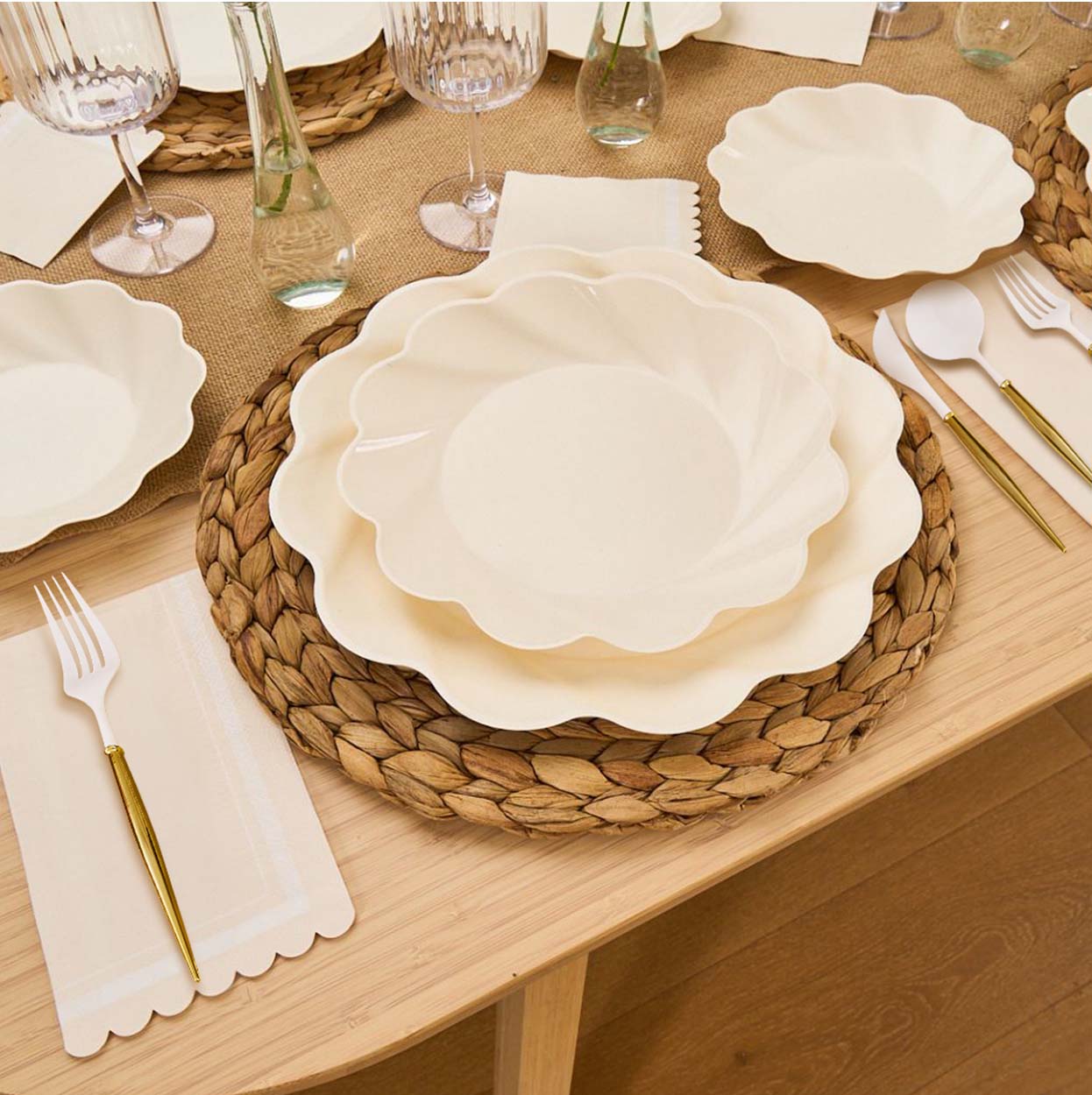 Create a stylish tablescape with Sophistiplate’s Eco-Friendly Table Setting- Cream, featuring scalloped cream plates, gold and white cutlery, woven placemats, glasses, napkins, and a beige table runner on a wooden table.