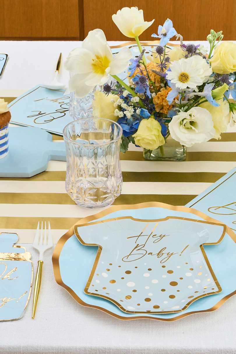 A baby shower table setting with gold and white striped runner, blue and gold onesie-shaped plates that read Hey Baby!, a clear glass, white cutlery, and a pastel floral centerpiece.