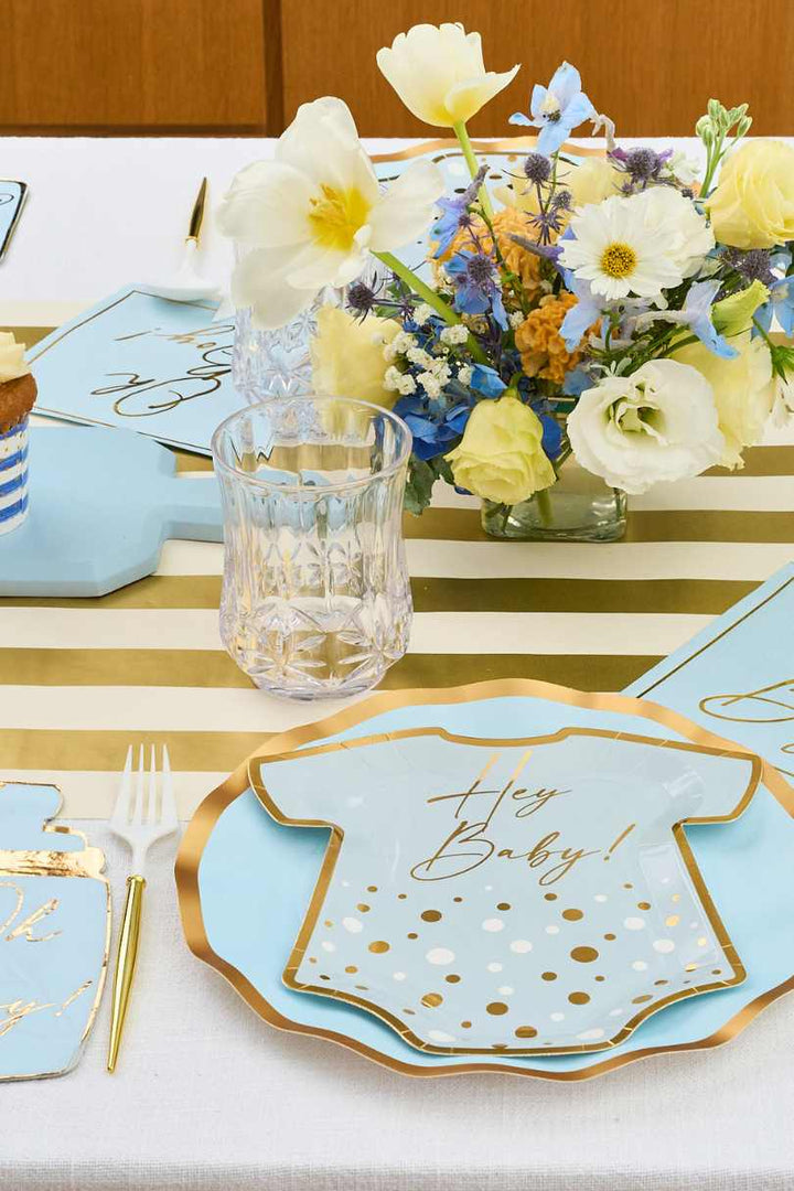 A baby shower table setting with gold and white striped runner, blue and gold onesie-shaped plates that read Hey Baby!, a clear glass, white cutlery, and a pastel floral centerpiece.