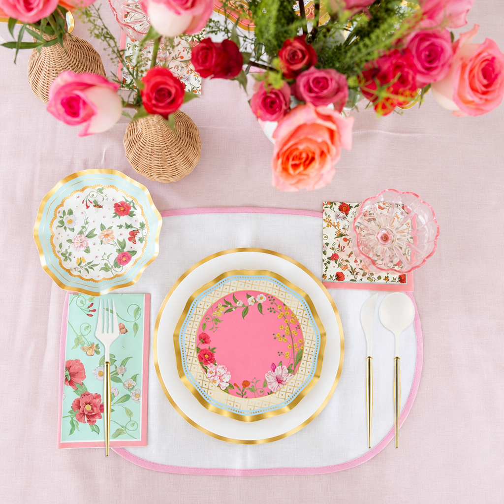Experience an elegant party-in-a-box with the Ivory Rococo Table Setting by Sophistiplate—designer disposable plates with intricate floral motifs, a pink placemat, coordinating floral napkins, chopsticks, a glass, two white spoons, and a bouquet of pink and orange roses in woven vases atop a light pink tablecloth.