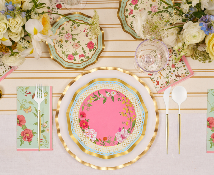 Experience the Ivory Rococo Table Setting by Sophistiplate, featuring designer disposable plates in pink and gold, a gold-rimmed white plate, floral napkins, elegant white cutlery, a wine glass, and sophisticated floral arrangements atop a cream-striped tablecloth.