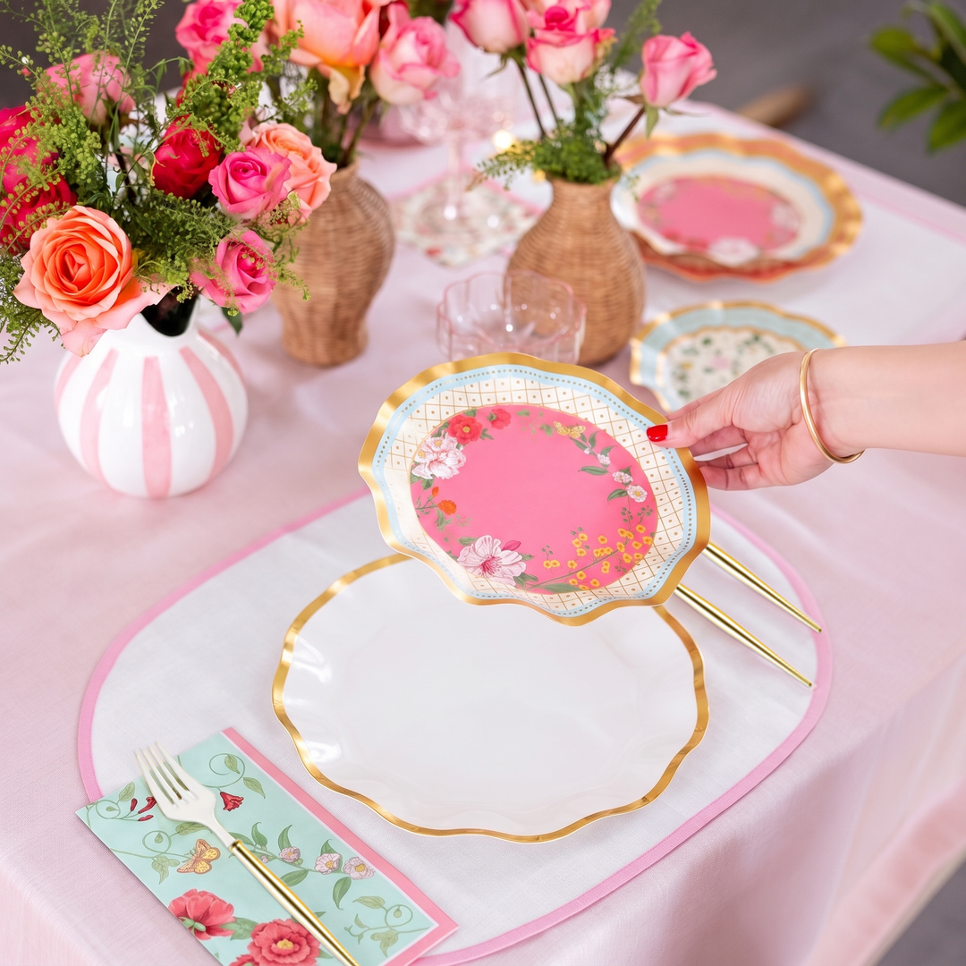 A hand places an Ivory Rococo Table Setting designer disposable plate by Sophistiplate on a white plate atop a set table with gold utensils, floral napkins, and vases of pink and orange roses on a pink tablecloth.