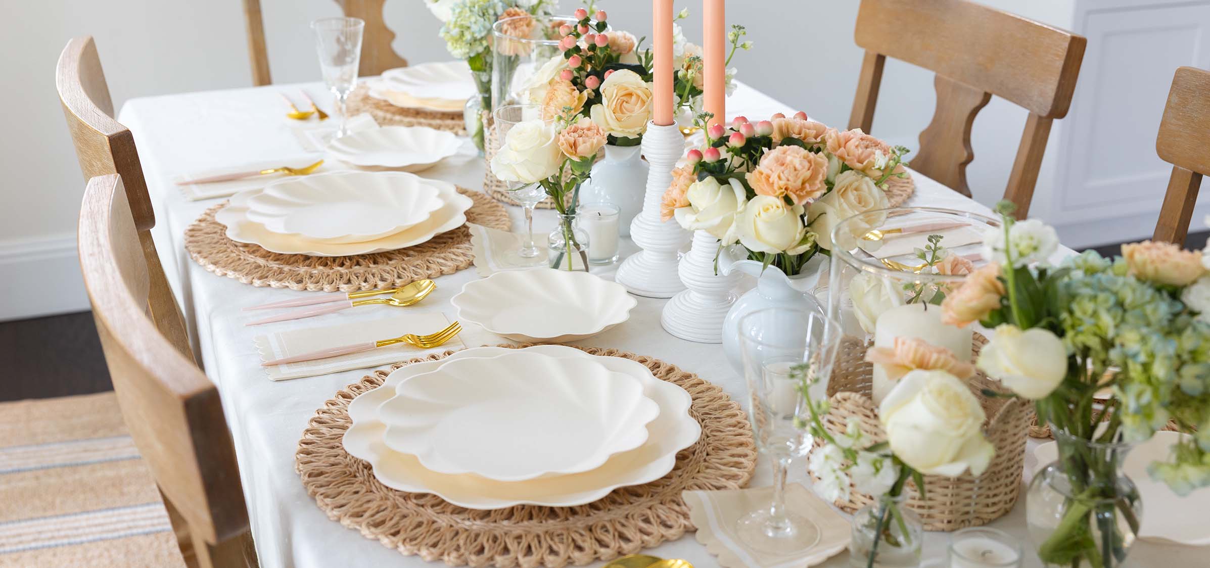 A dining table set for a meal with scalloped white plates on woven placemats, gold cutlery, and pastel floral centerpieces, surrounded by wooden chairs and white taper candles in holders.