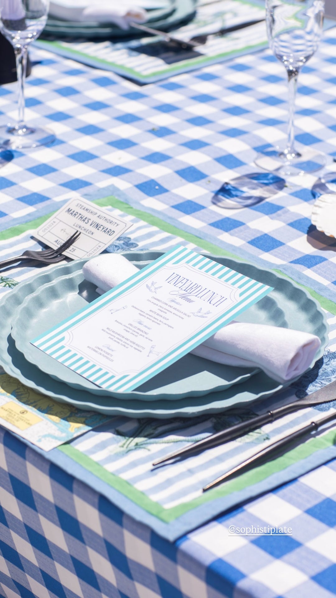 A close-up of a blue and white checkered tablecloth with a place setting, featuring an eco-friendly green charger, Sophistiplate Simply Eco Dinner Plate - Pool, bamboo fiber plates, menu, cutlery, glassware, and a rolled white napkin in sunlight.