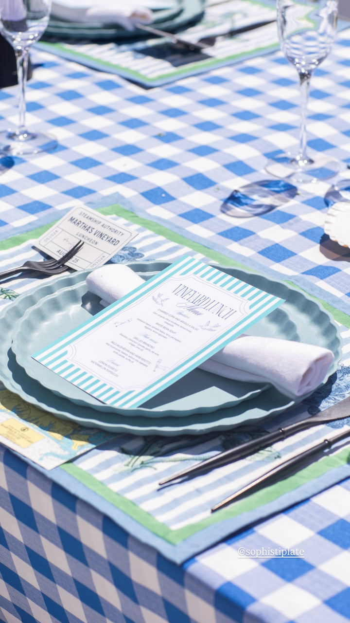 A close-up of a blue and white checkered tablecloth with a place setting, featuring an eco-friendly green charger, Sophistiplate Simply Eco Dinner Plate - Pool, bamboo fiber plates, menu, cutlery, glassware, and a rolled white napkin in sunlight.