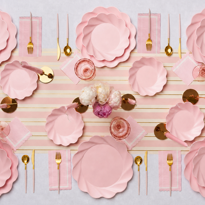 A table set with the Eco-Friendly Table Setting- Blush by Sophistiplate LLC, featuring compostable party plates with pink scalloped edges, gold flatware, pink napkins, pink glasses, and a centerpiece of white and pink flowers atop a striped table runner.