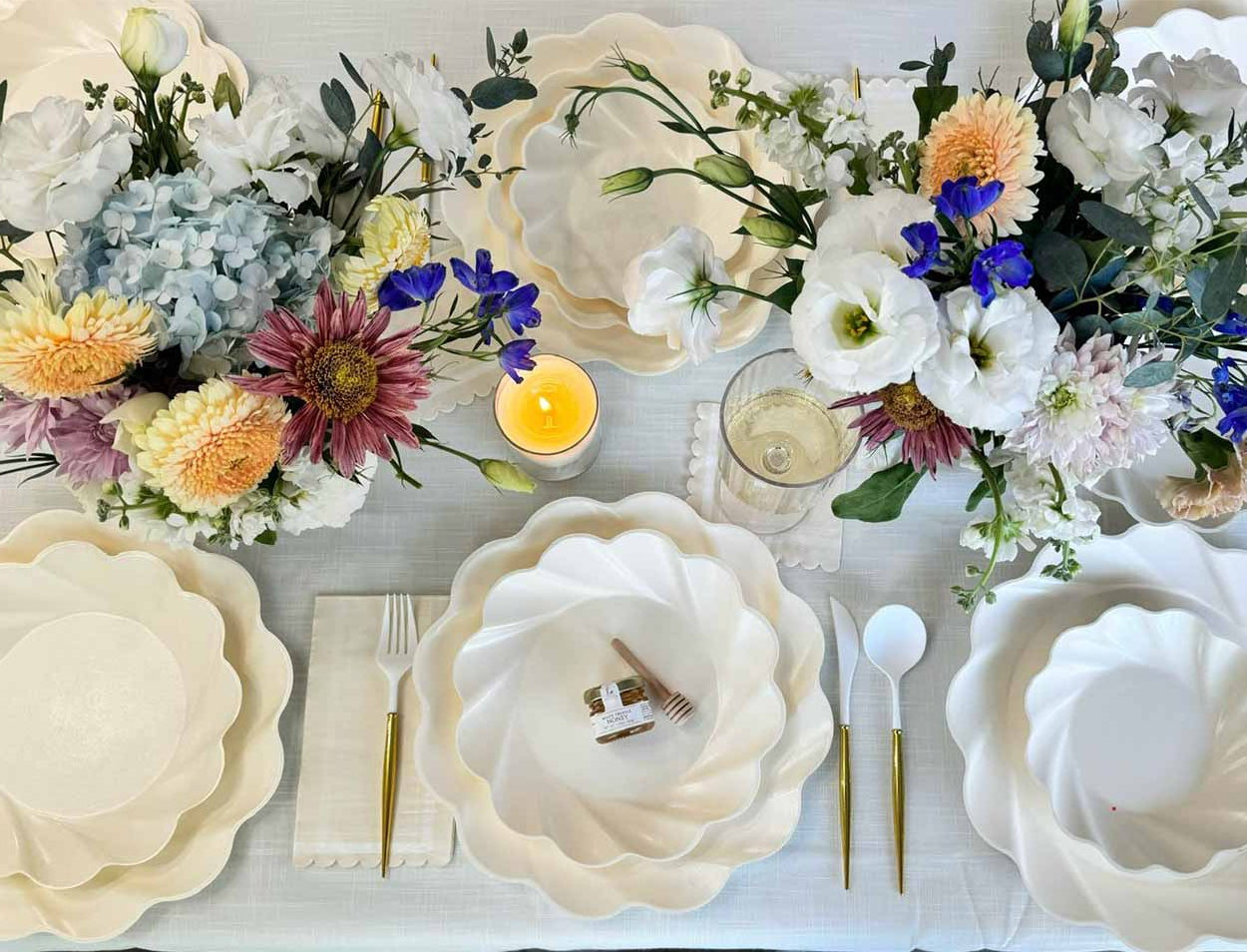 A stunning table featuring Sophistiplate’s Eco-Friendly Table Setting—cream scalloped compostable plates, gold cutlery, white napkins, floral centerpieces, a candle, wine glass, and a honey jar with dipper.