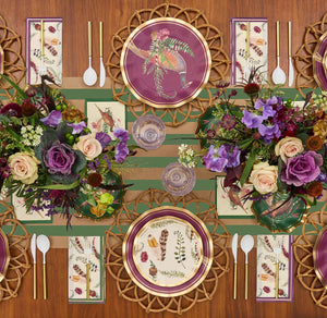 The Feather Grove Table Setting by Sophistiplate features elegant purple and gold disposable plates, botanical napkins, gold utensils, and white-handled cutlery on a wooden table with floral accents and a green-striped runner for a stunning fall look.