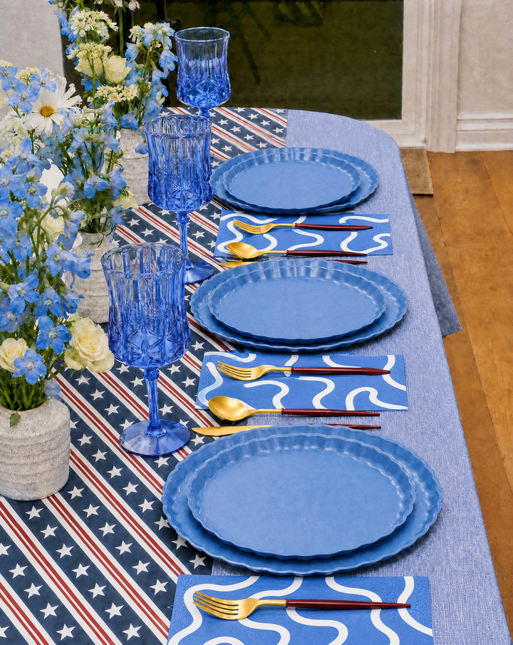 The 4th of July Table for 16+ in French Blue by Sophistiplate LLC features compostable bamboo plates, gold utensils, blue patterned napkins, blue glass goblets, vases with white and blue flowers, and a star-spangled wavy-striped runner.
