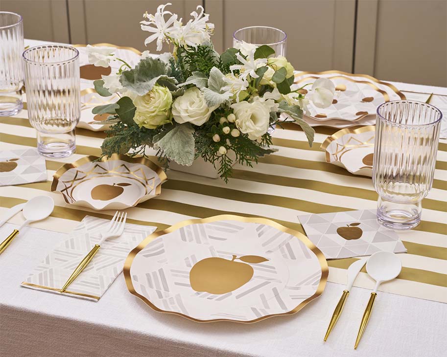 Create an elegant Rosh Hashanah table setting with the “Rosh Tablescape for 16 Guests: Golden Apple” by Sophistiplate LLC, featuring designer disposable plates adorned with gold-rimmed edges and golden apple motifs, complemented by clear ribbed glasses, gold-and-white cutlery, and a white and green floral centerpiece arranged on a gold-striped table runner.