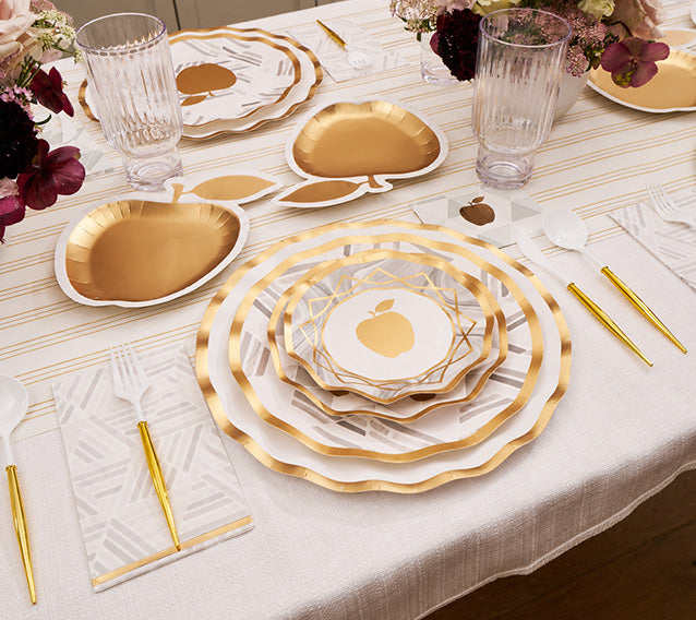 Create a stunning Rosh Hashanah table with the Sophistiplate LLC “Rosh Tablescape for 16 Guests: Golden Apple,” featuring gold and white designer disposable plates with gold apple motifs, clear glasses, gold utensils, and floral centerpieces on a cream-striped tablecloth.