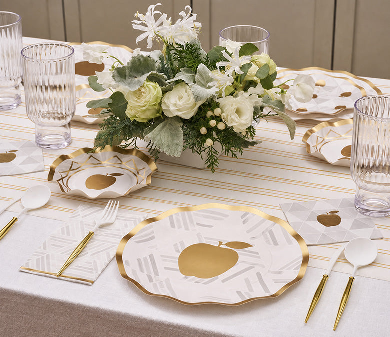 Create an elevated Rosh Hashanah table with the Rosh Tablescape for 16 Guests: Golden Apple by Sophistiplate LLC, featuring designer disposable plates adorned with a gold apple motif, gold utensils, clear ribbed glasses, and a white-green floral centerpiece arranged atop a white tablecloth and striped runner.