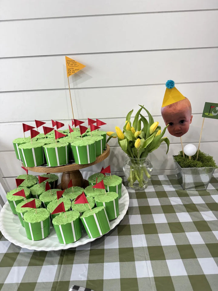 Nestled in Simply Baked Large Paper Baking Cups | Green Vertical, green cupcakes with red flags sit on a stand, accompanied by yellow tulips in a vase and a playful decor piece featuring a baby doll head with a party hat, golf ball, and flag—all beautifully arranged atop a green-checked tablecloth.