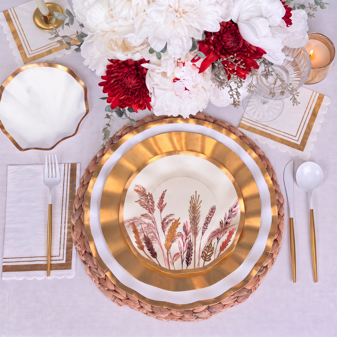 The Harvest Gold Table Setting by Sophistiplate features gold and white decorative botanical paper plates, gold flatware, a woven placemat, a white napkin, and a floral centerpiece with white and red blooms atop a crisp white tablecloth, creating an effortless tablescape in a box.