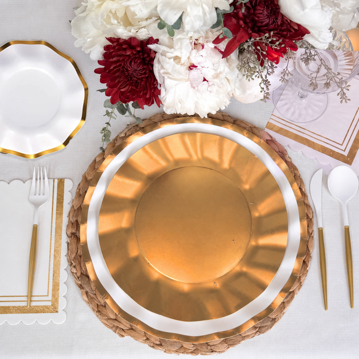 Create an effortlessly beautiful tablescape with the Harvest Gold Table Setting by Sophistiplate—this box includes a gold plate on a woven mat, decorative paper plates, gold and white utensils, scalloped napkins, and a floral centerpiece with white and red flowers atop a crisp white tablecloth.
