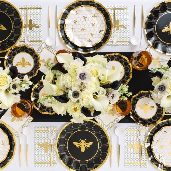 A formal table setting with black, white, and gold bee-themed plates, gold cutlery, amber glasses, and floral centerpieces featuring white blooms, arranged neatly on a patterned tablecloth.