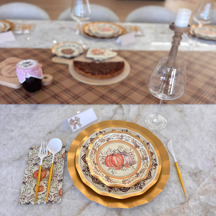 A Turkey & Gourds: Thanksgiving Table for 16 setting with heavy-duty paper plates featuring a gold-rimmed pumpkin design, white and gold cutlery, and a pumpkin-print napkin arranged on a marble table with a plaid runner and dessert centerpiece.