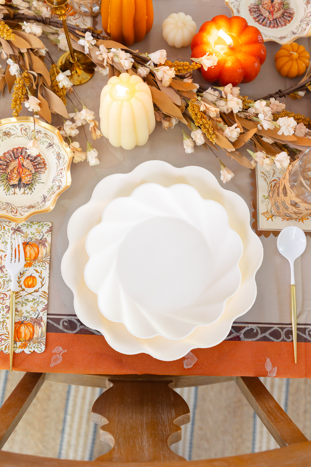 The Ivory Harvest Table Setting by Sophistiplate creates a beautiful Thanksgiving tablescape with scalloped white plates, pumpkin decorations, candles, floral garland, and autumn-themed napkins and cutlery on a festive tablecloth. This thoughtfully curated Thanksgiving tableware set features eco-friendly disposable plates that blend elegance with sustainability for your holiday gathering.