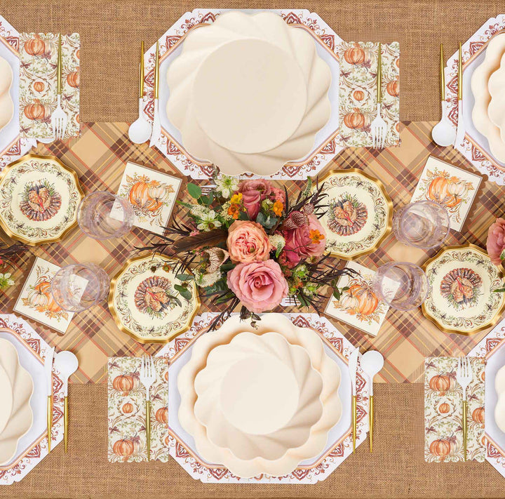 Enjoy a festive tablescape with the Sophistiplate Ivory Harvest Table Setting, featuring floral centerpieces, eco-friendly disposable plates adorned with pumpkin illustrations, a plaid table runner, gold utensils, glasses, and napkins—all elegantly arranged on a burlap tablecloth.