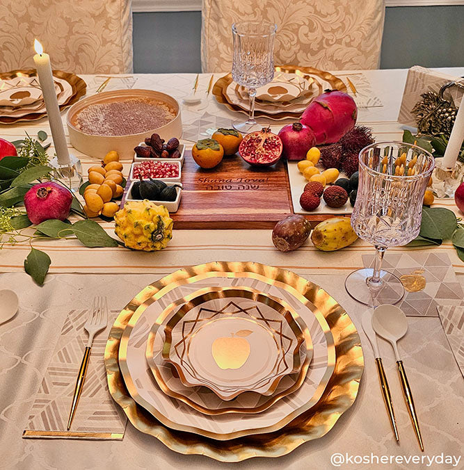 An elegant table setting features gold-trimmed plates, crystal glasses, candles, assorted fruits, nuts, and honey arranged on a decorative tablecloth. A wooden cutting board with sliced bread sits at the center, highlighted by beautifully designed Golden Apple Paper Cocktail Napkins from Sophistiplate for a festive Rosh Hashanah touch.