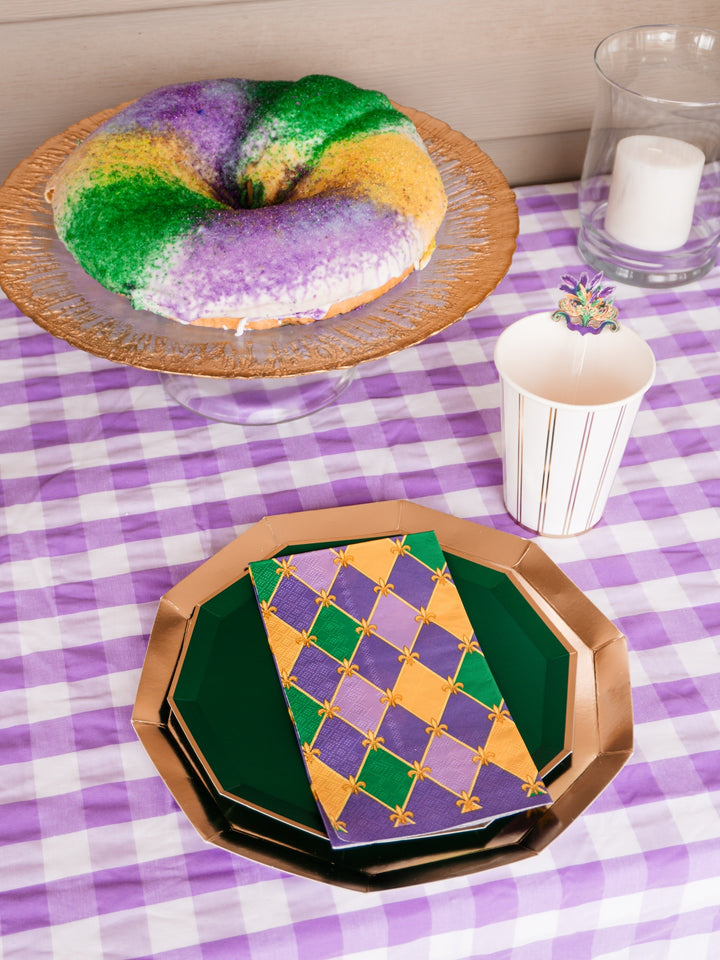 A Sophistiplate Let’s Mardi Table Setting features a King Cake topped with green, yellow, and purple sugar, arranged on a purple checkered tablecloth along with a candle, festive cup, Mardi Gras party plates, and a napkin.