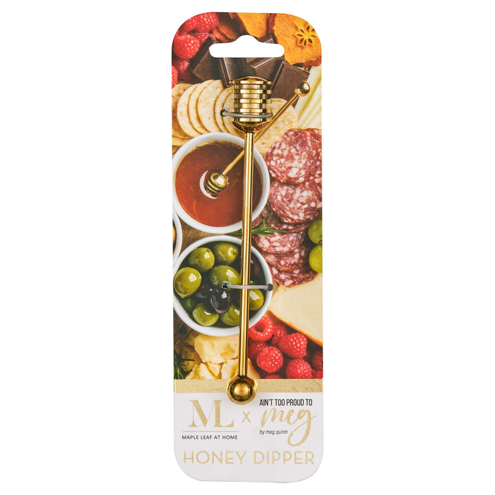 The Maple Meg Quinn Gold Stainless Steel Honey Dipper is packaged on a card with a charcuterie board image and meats, olives, fruit, and honey. The label says “ML x Meg Honey Dipper.”.