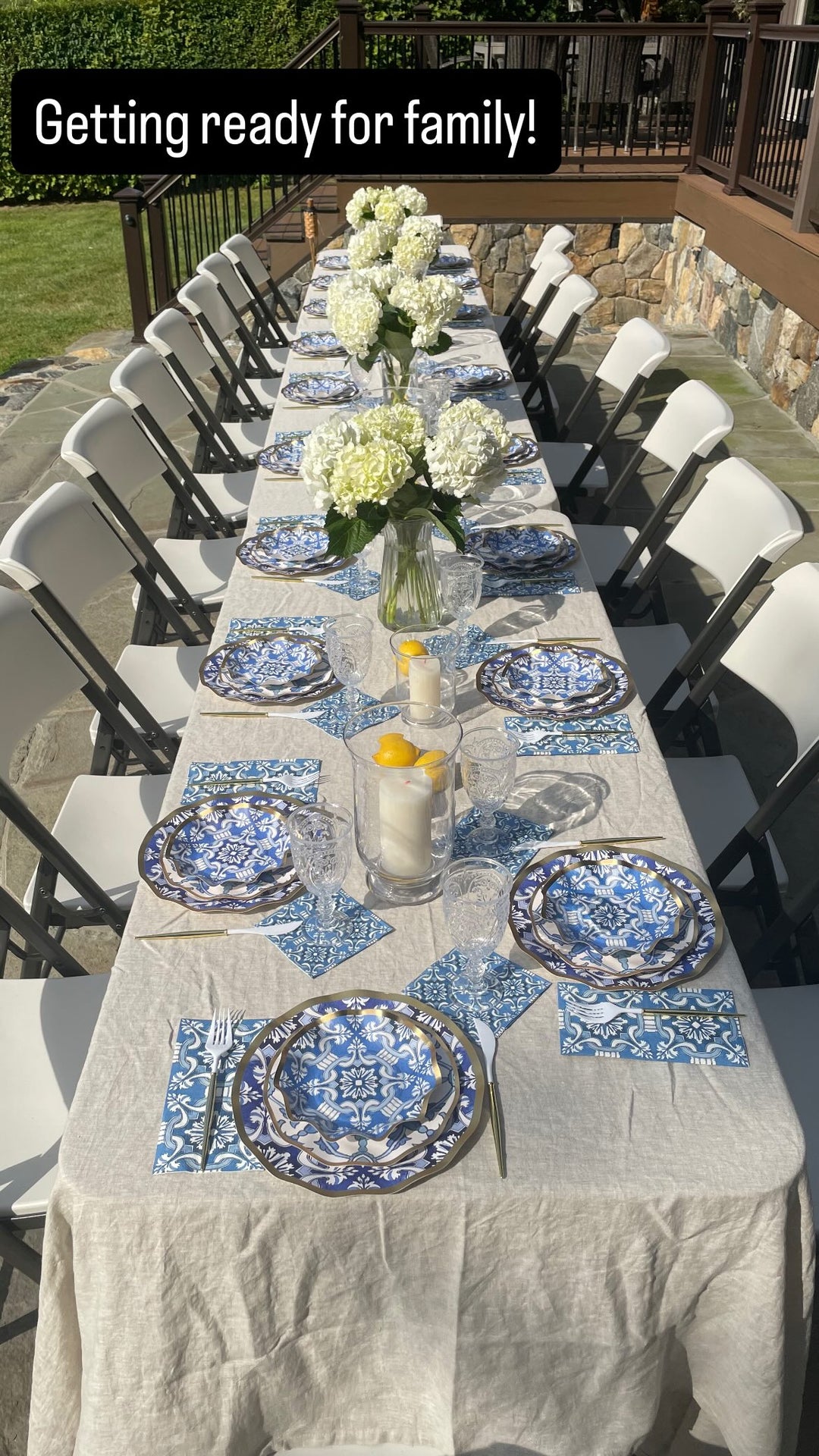 A long outdoor table set for a gathering features Sophistiplate LLC’s Moroccan Table Setting, with blue-patterned heavy duty paper plates, matching napkins, clear glasses, white flowers in vases, and a candle centerpiece. Folding chairs line both sides under sunny weather—bringing the feel of a party-in-a-box to life.