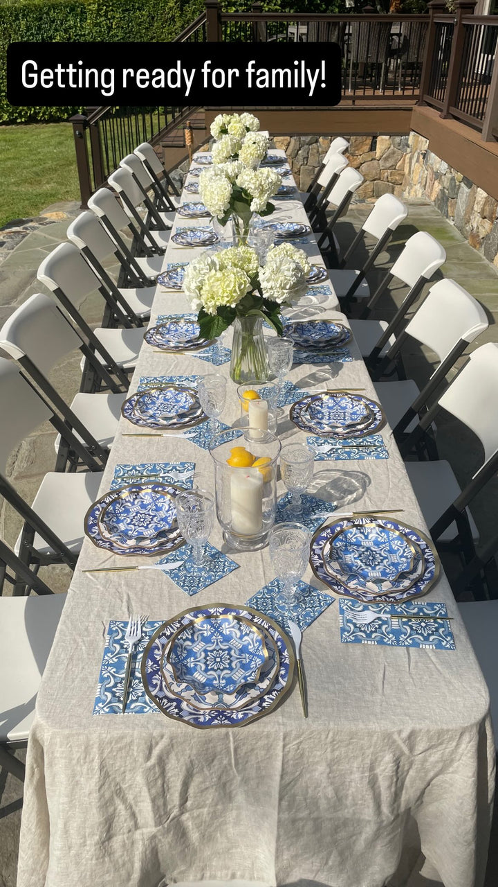 A long outdoor table set for a gathering features Sophistiplate LLC’s Moroccan Table Setting, with blue-patterned heavy duty paper plates, matching napkins, clear glasses, white flowers in vases, and a candle centerpiece. Folding chairs line both sides under sunny weather—bringing the feel of a party-in-a-box to life.